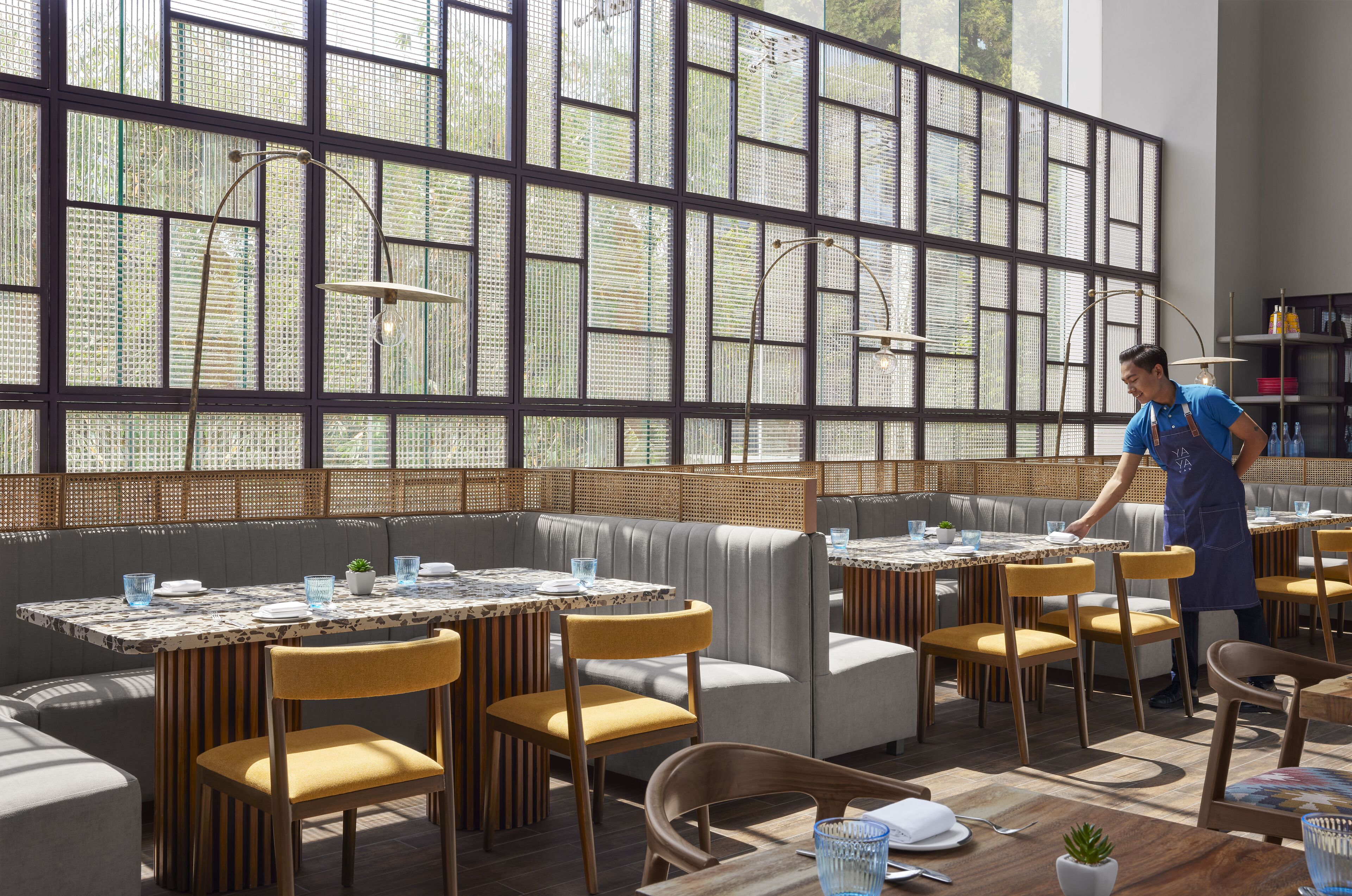 A restaurant with high windows, cushioned seating, and a waiter attending to a table.