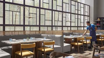 A restaurant with high windows, cushioned seating, and a waiter attending to a table.