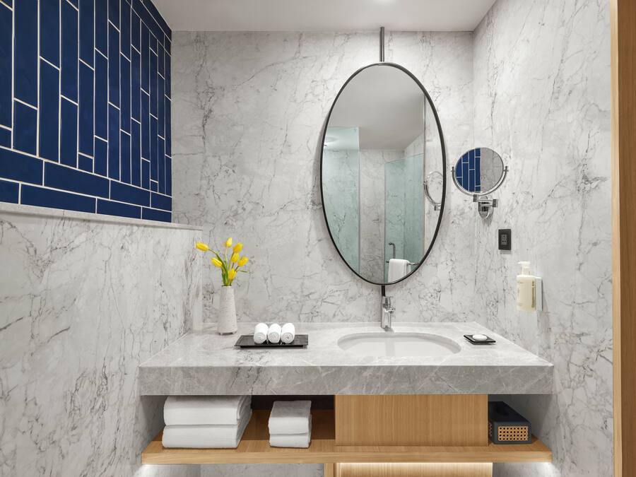 Un baño moderno con un lavabo de mármol, un espejo ovalado grande y un jarrón pequeño con flores amarillas.