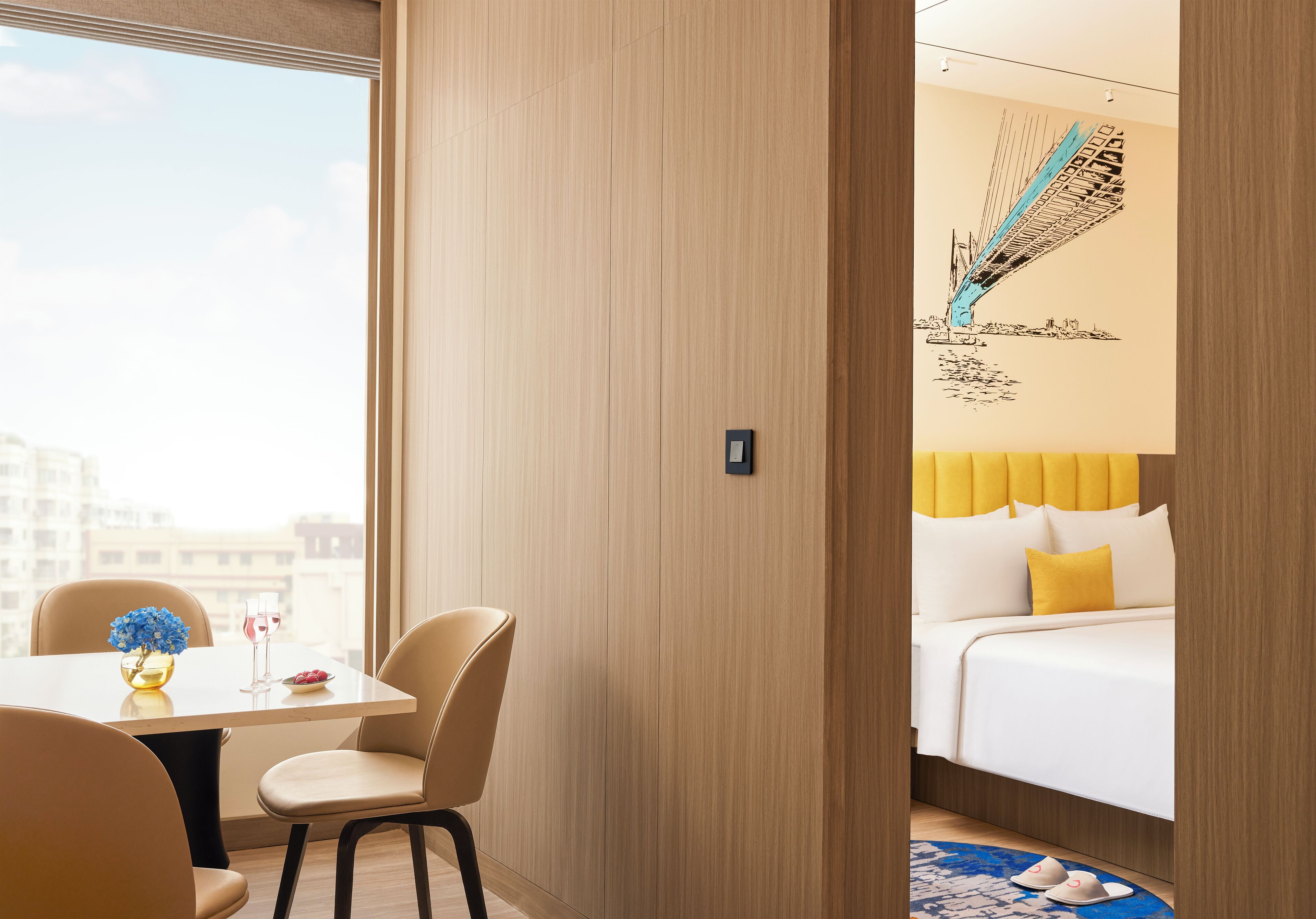 A hotel room with a bed, a dining area with a table and chairs, and a view of the city through large windows.