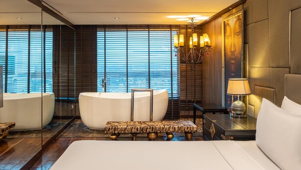 Een moderne hotelkamer met een groot bed, een badkuip, een bankje, een nachtkastje met een lamp, en uitzicht op de stad door grote ramen.