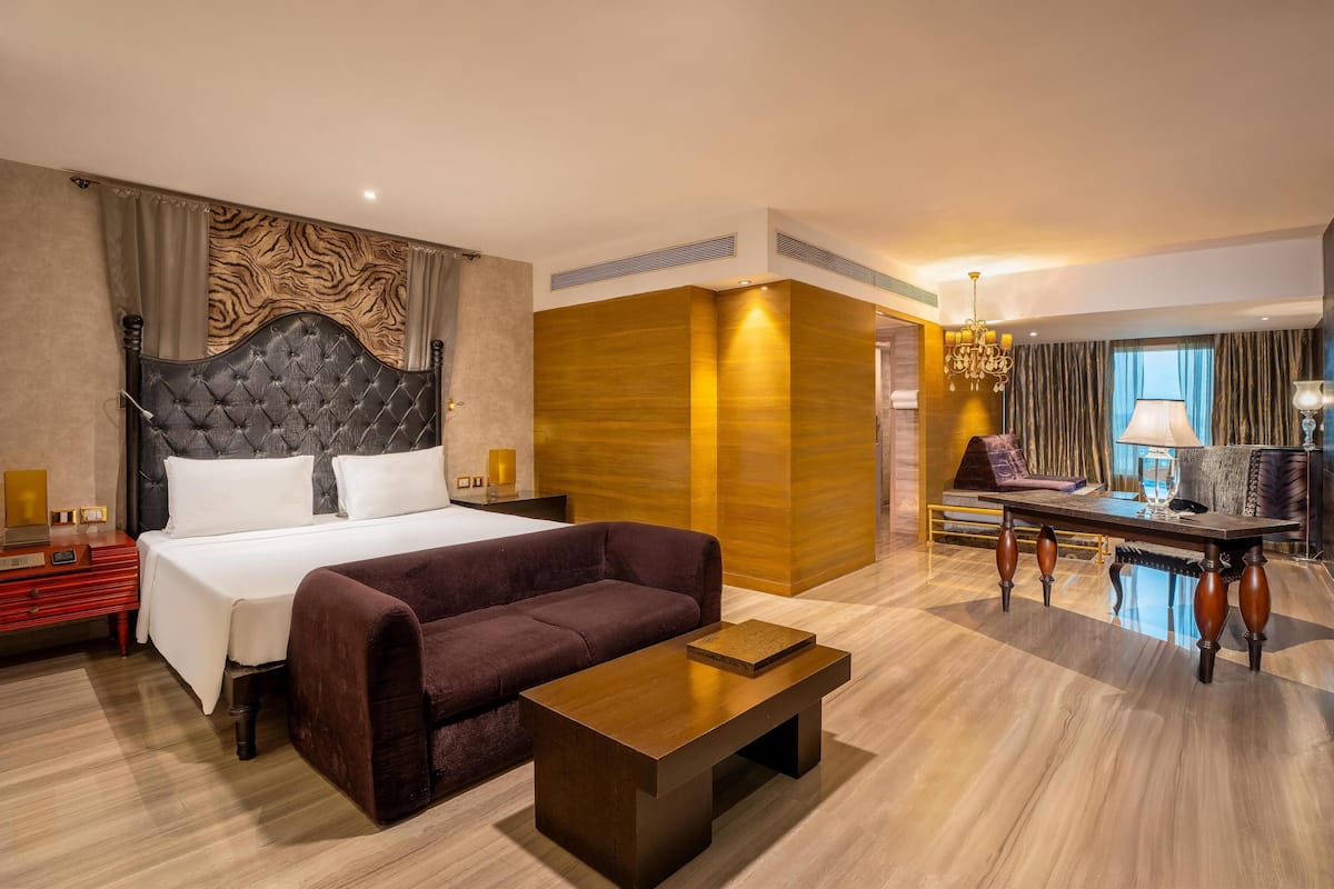 Une chambre d’hôtel avec un grand lit, un canapé, une table basse en bois, un bureau avec une lampe et une vue sur l’extérieur.