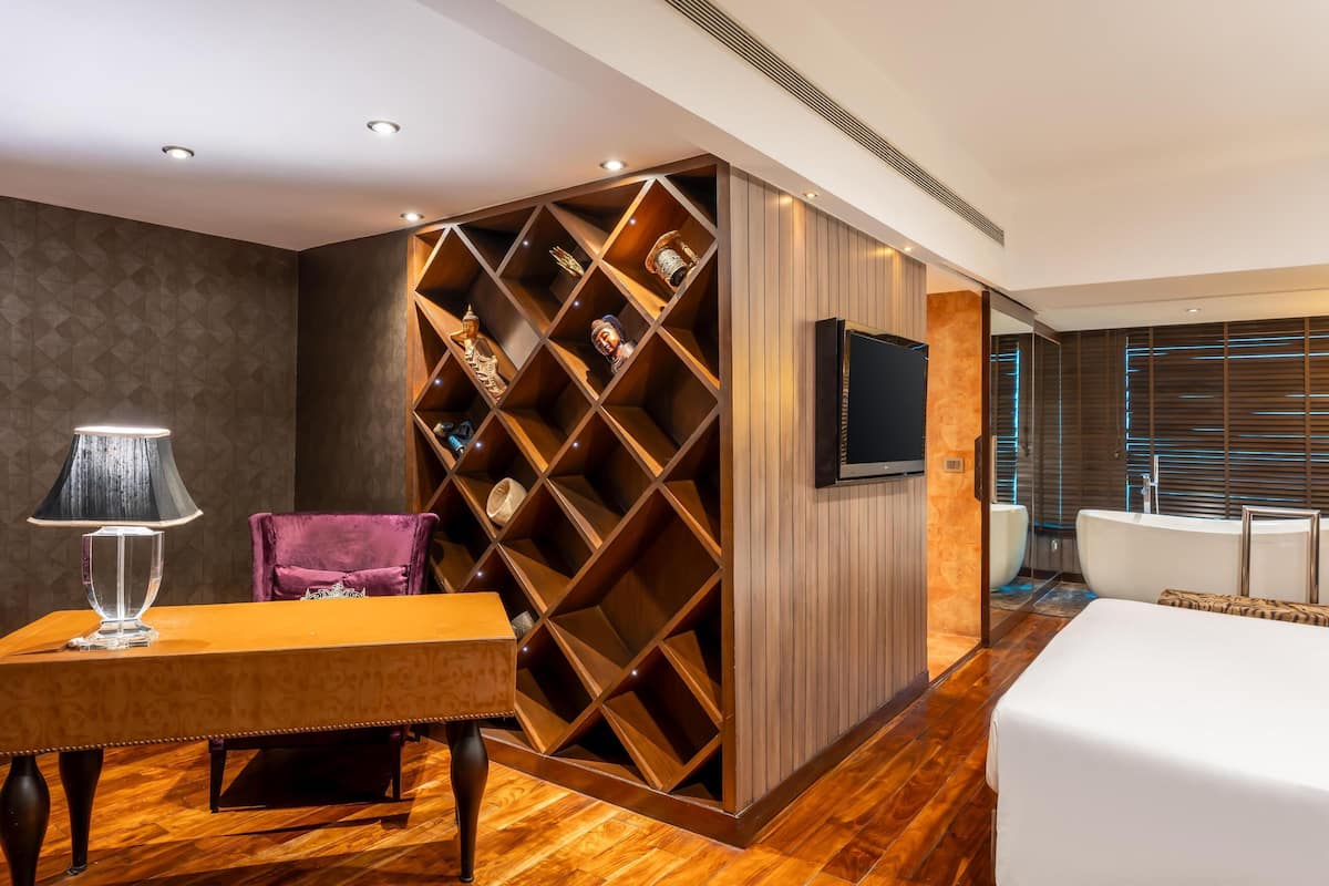 Une chambre d’hôtel moderne avec un plancher en bois, un grand lit, un téléviseur fixé au mur, une bibliothèque intégrée en bois agrémentée d’objets décoratifs et un coin salon avec un fauteuil violet.