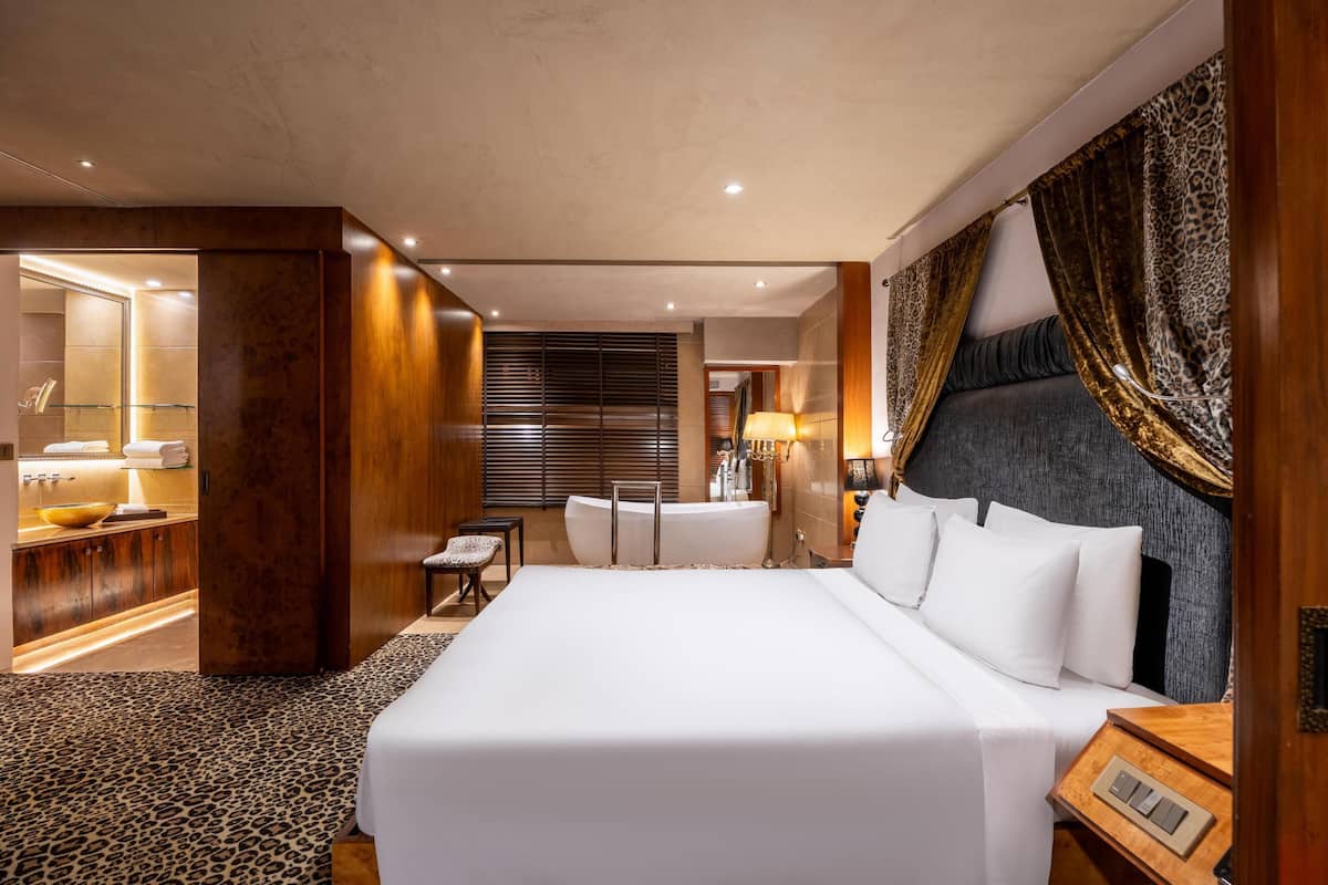 Une chambre d’hôtel moderne dotée d’un grand lit, d’un coin salon avec un fauteuil et une table, d’une baignoire et d’une salle de bain avec une douche à parois de verre.