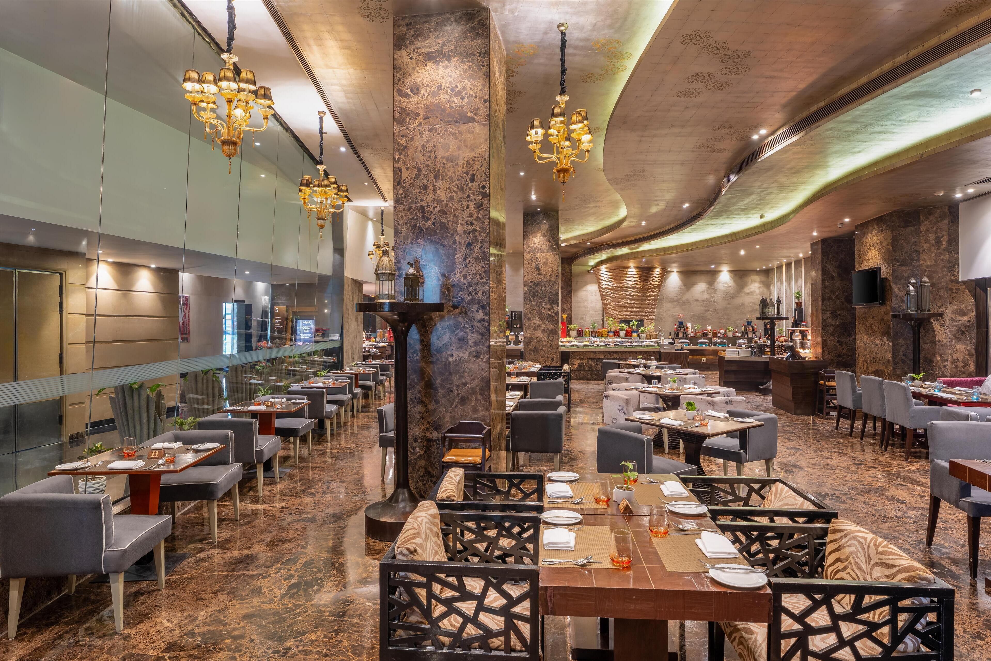 A spacious restaurant with high ceilings, chandeliers, and a central column. Tables are arranged for dining, and there are various seating options.