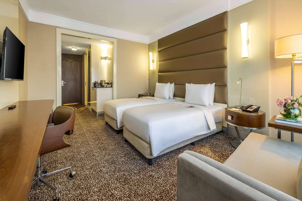 Ein Hotelzimmer mit zwei Betten, einem Schreibtisch, einem Sessel, einem Fernseher und einer Lampe.