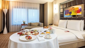 Ein Hotelzimmer mit Esstisch, gedecktem für das Frühstück, einer Couch, einem Bett und einem großen Fenster mit Vorhängen.