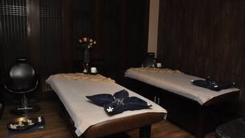 Relax in a serene space with two massage tables, a small table with a vase and candle, and a comfortable chair.