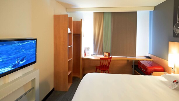 Habitación de hotel con cama, escritorio, televisión, armario y ventana con cortinas.