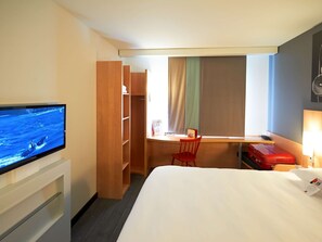Habitación de hotel con cama, escritorio, televisión, armario y ventana con cortinas.