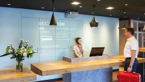 Un mostrador de recepción con una pantalla digital, un jarrón de flores y una maleta roja.