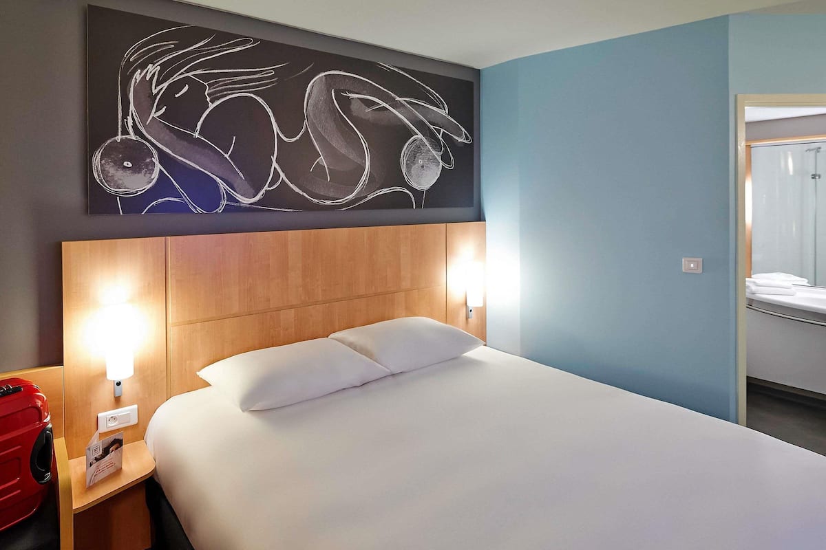 A hotel room with a large bed, two bedside lamps, a wooden headboard, a wall art piece, and a red suitcase on the left side.