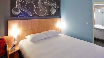 A hotel room with a large bed, two bedside lamps, a wooden headboard, a wall art piece, and a red suitcase on the left side.