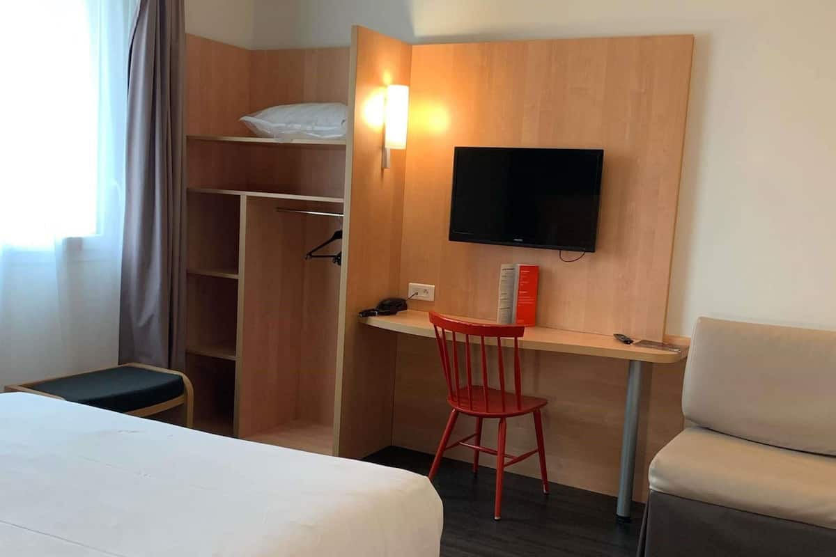 A hotel room with a bed, a desk with a chair, a TV mounted on the wall, and a wardrobe.