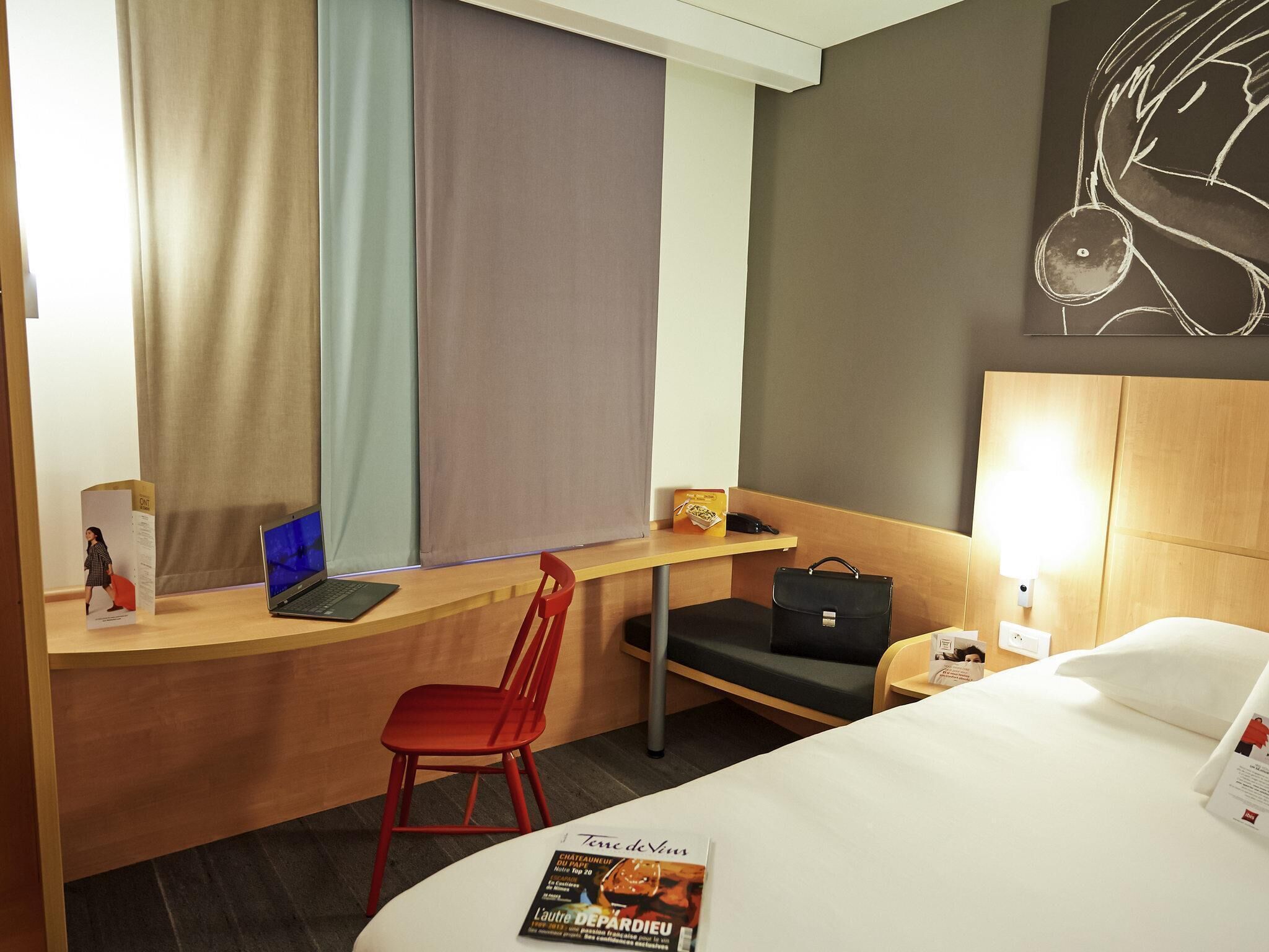 Une chambre d’hôtel avec un lit, un bureau avec un ordinateur portable, une chaise rouge, un sac noir, une horloge murale et un magazine sur le lit.