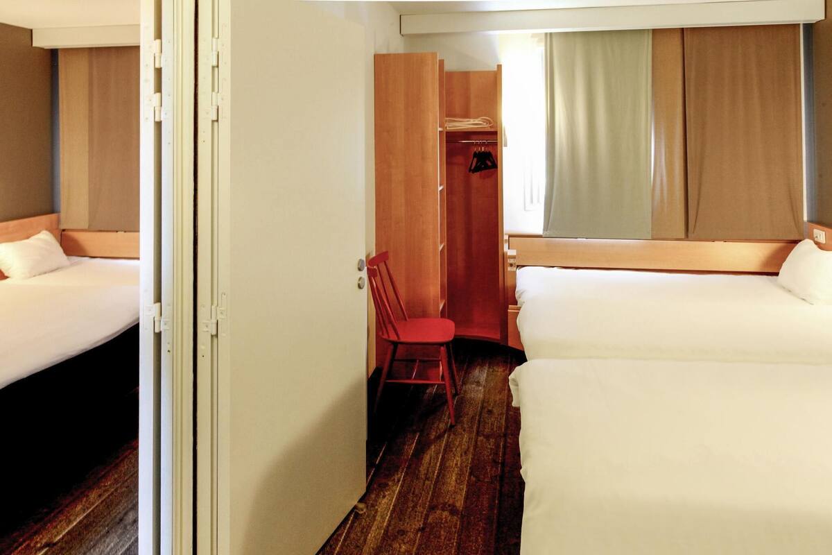 Una habitación de hotel con dos camas, un armario de madera, una silla roja y una ventana con cortinas.