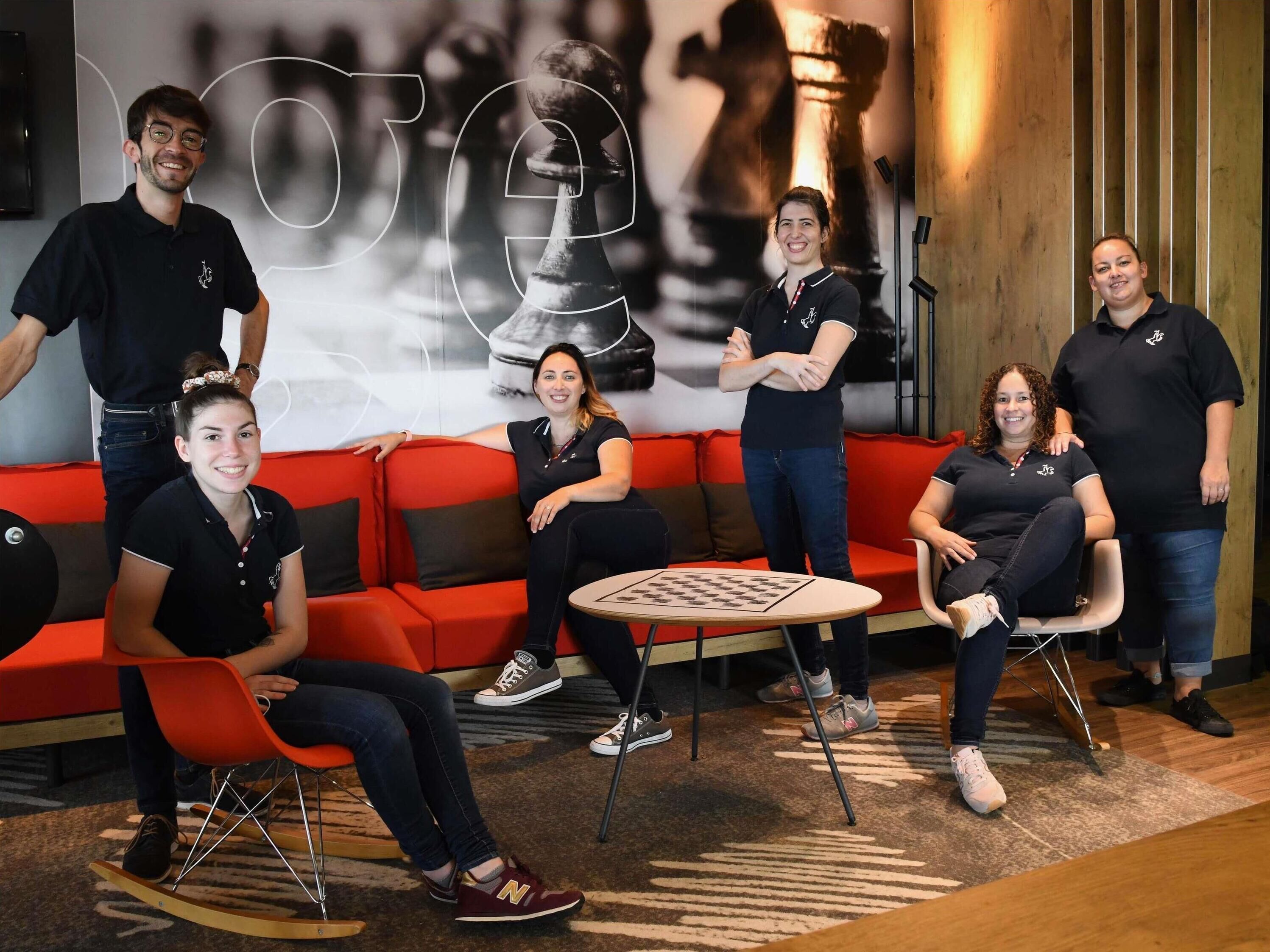 A group of people in a modern lounge area with a chess table, red seating, and a large abstract wall art piece.