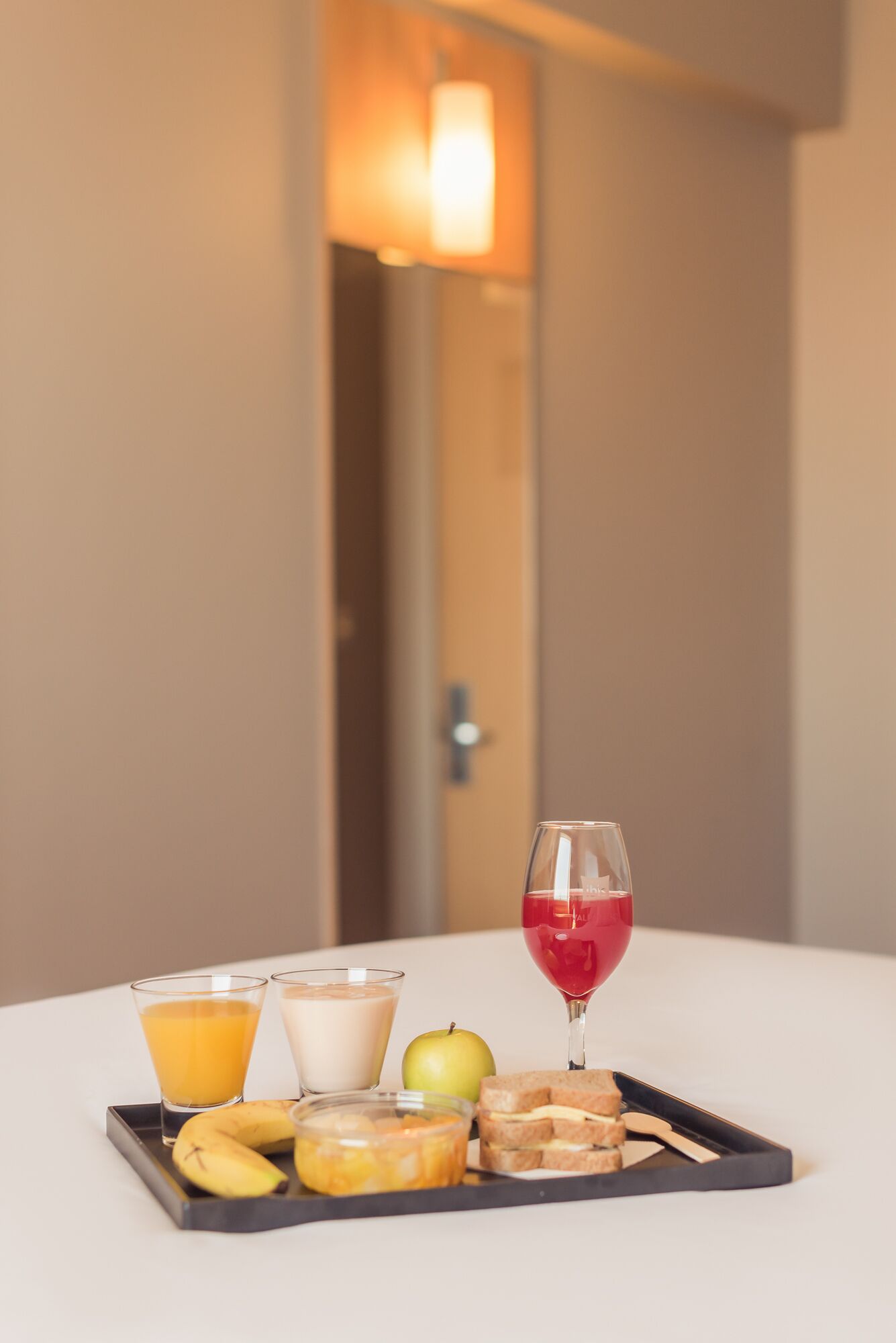 En bakke med morgenmadsprodukter, herunder juice, mælk, en rød drik, en banan, et æble og en sandwich på et hvidt bord.