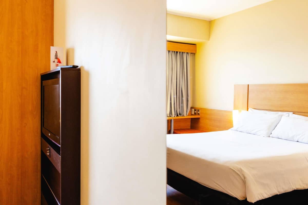 A hotel room with a bed, a wooden headboard, a television, and a curtain.