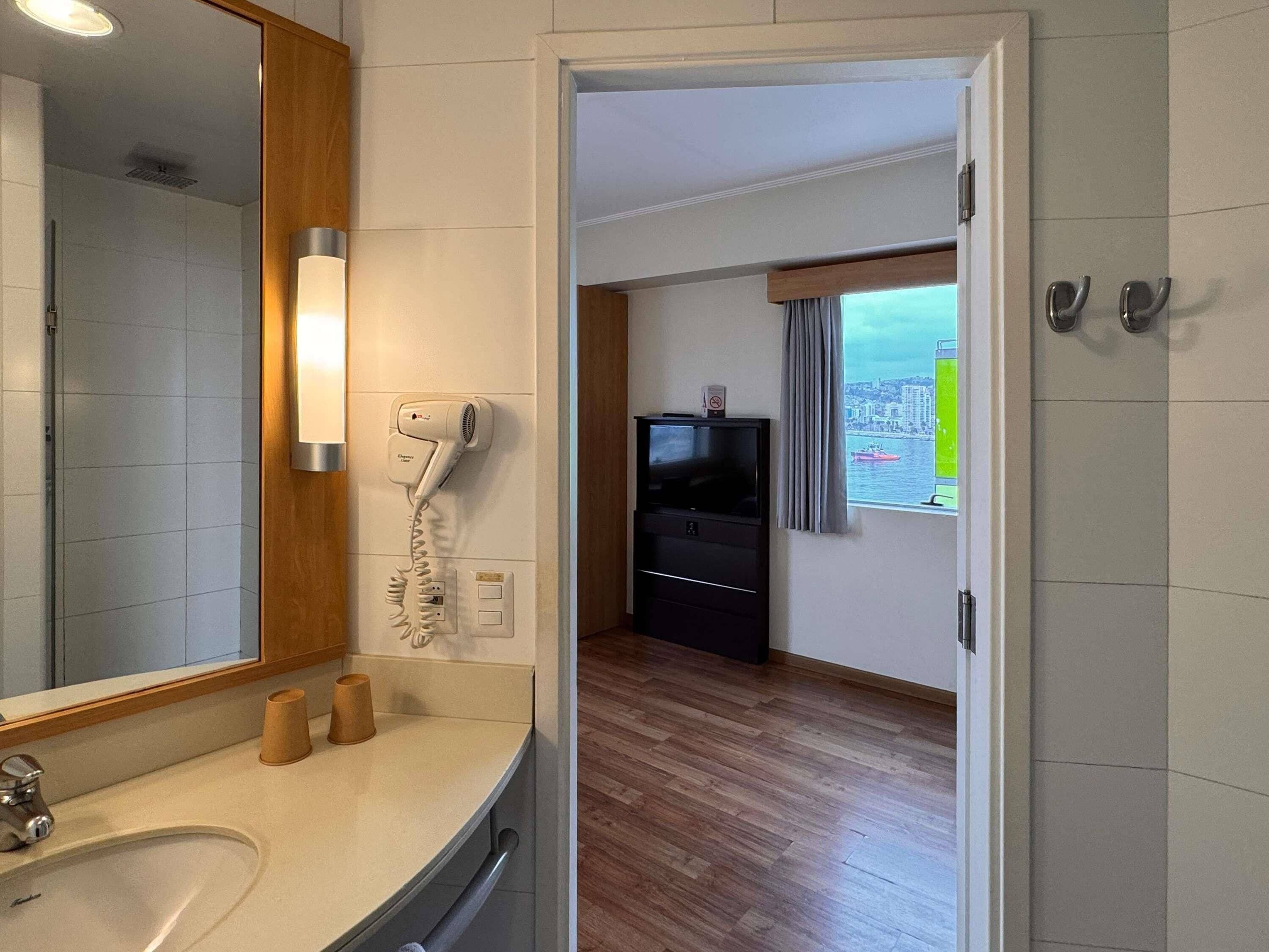 A hotel room with a bathroom, a hairdryer, a TV, and a window with a view of the city.