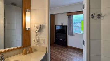 A hotel room with a bathroom, a hairdryer, a TV, and a window with a view of the city.