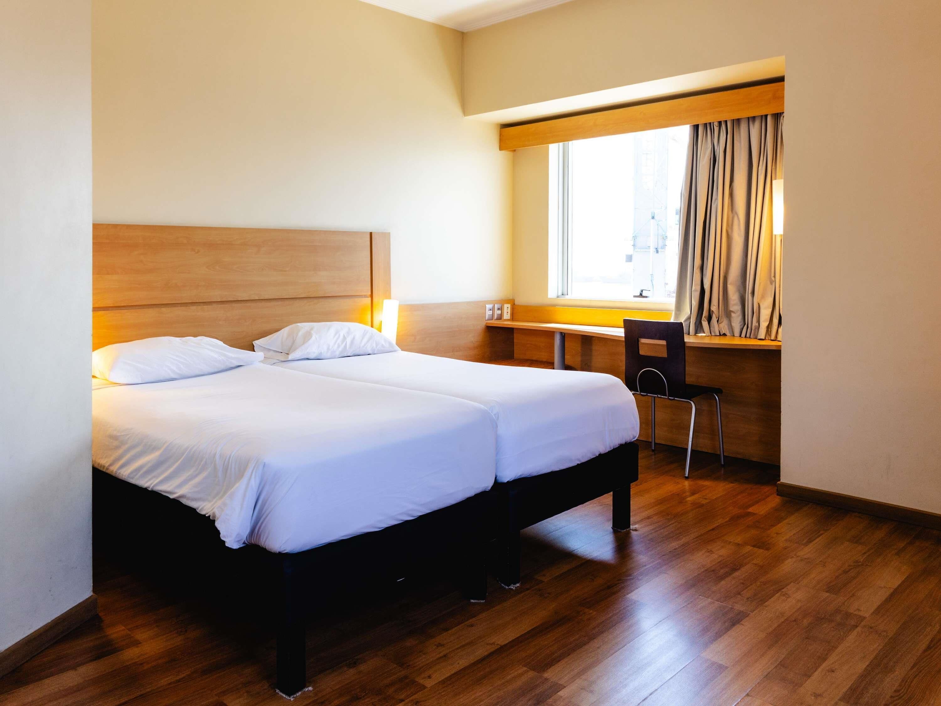 A hotel room with a double bed, a desk, a chair, and a window with curtains.