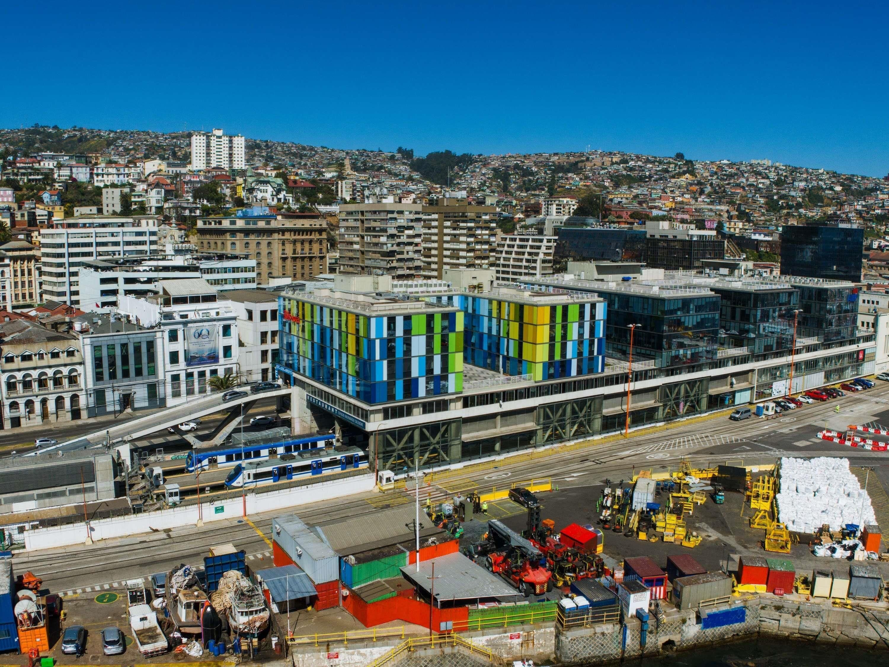 A modern building with a colorful facade, a train station in the foreground, and a densely populated hillside in the background.