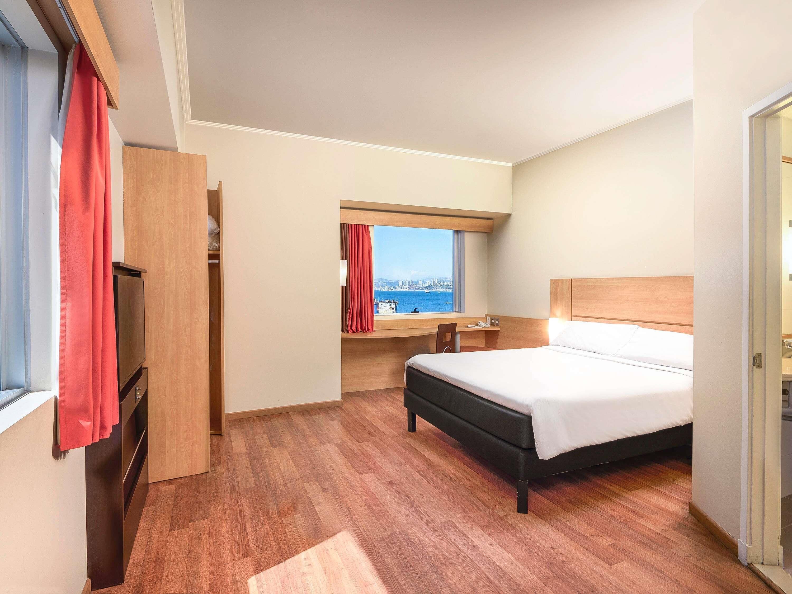 A hotel room with a bed, a desk, a window with a view of the sea, and a wardrobe.