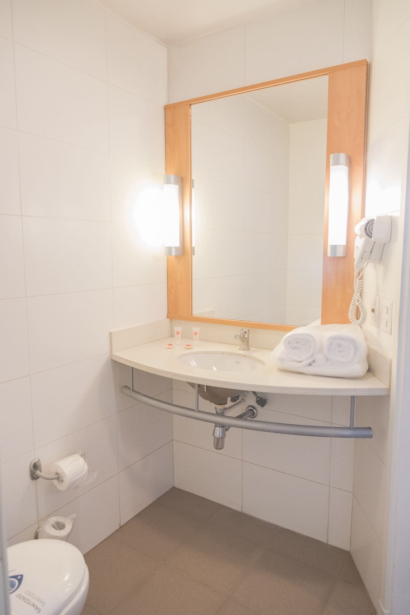A bathroom with a white sink, mirror, and towel rack. There is a toilet with a paper towel holder and a hand dryer.