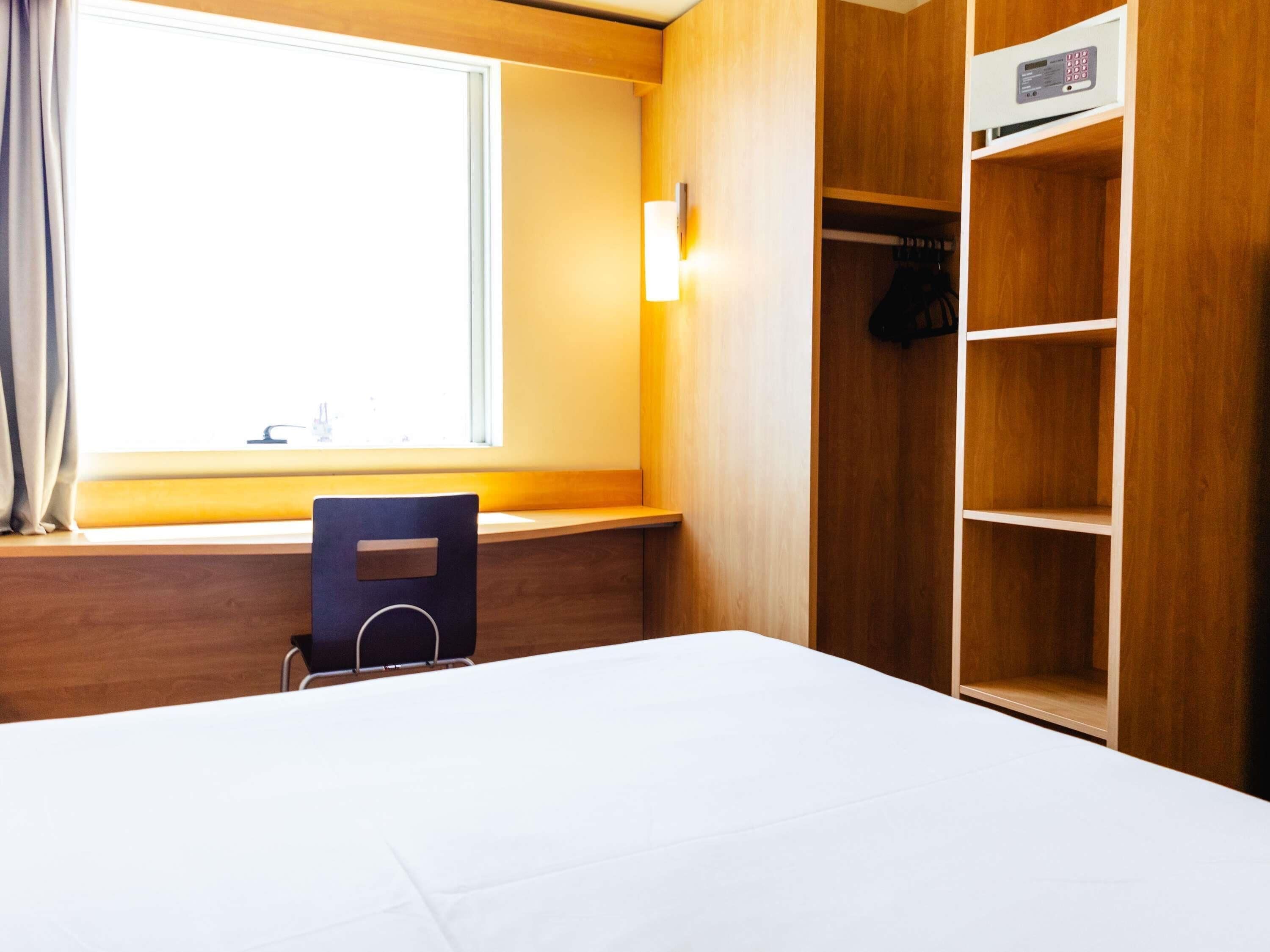 A hotel room with a bed, a desk, a chair, a window with curtains, and a wooden wardrobe with shelves.