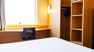 A hotel room with a bed, a desk, a chair, a window with curtains, and a wooden wardrobe with shelves.