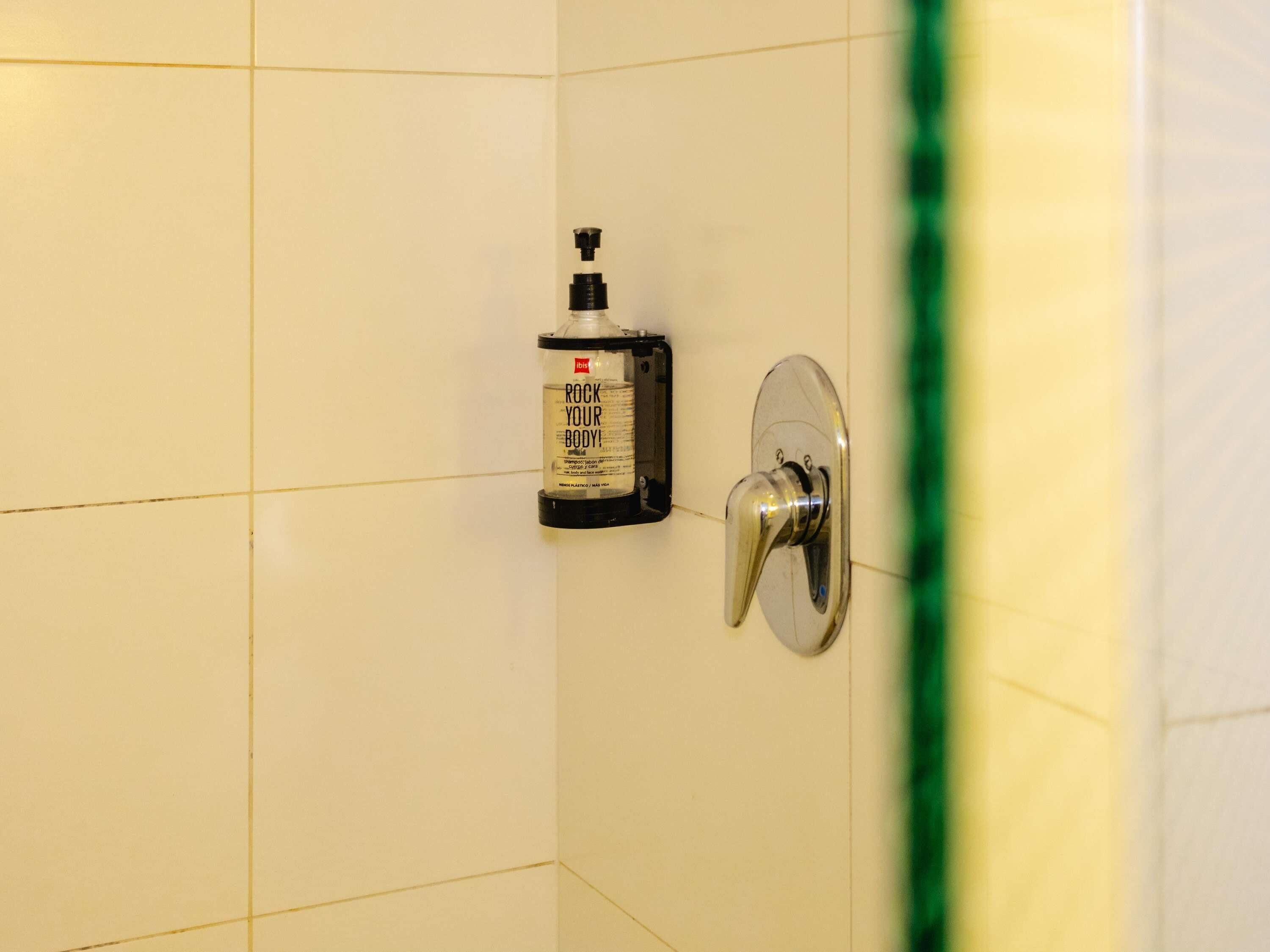 Un baño con paredes de azulejos blancos, un dispensador de jabón montado en la pared y un grifo cromado.