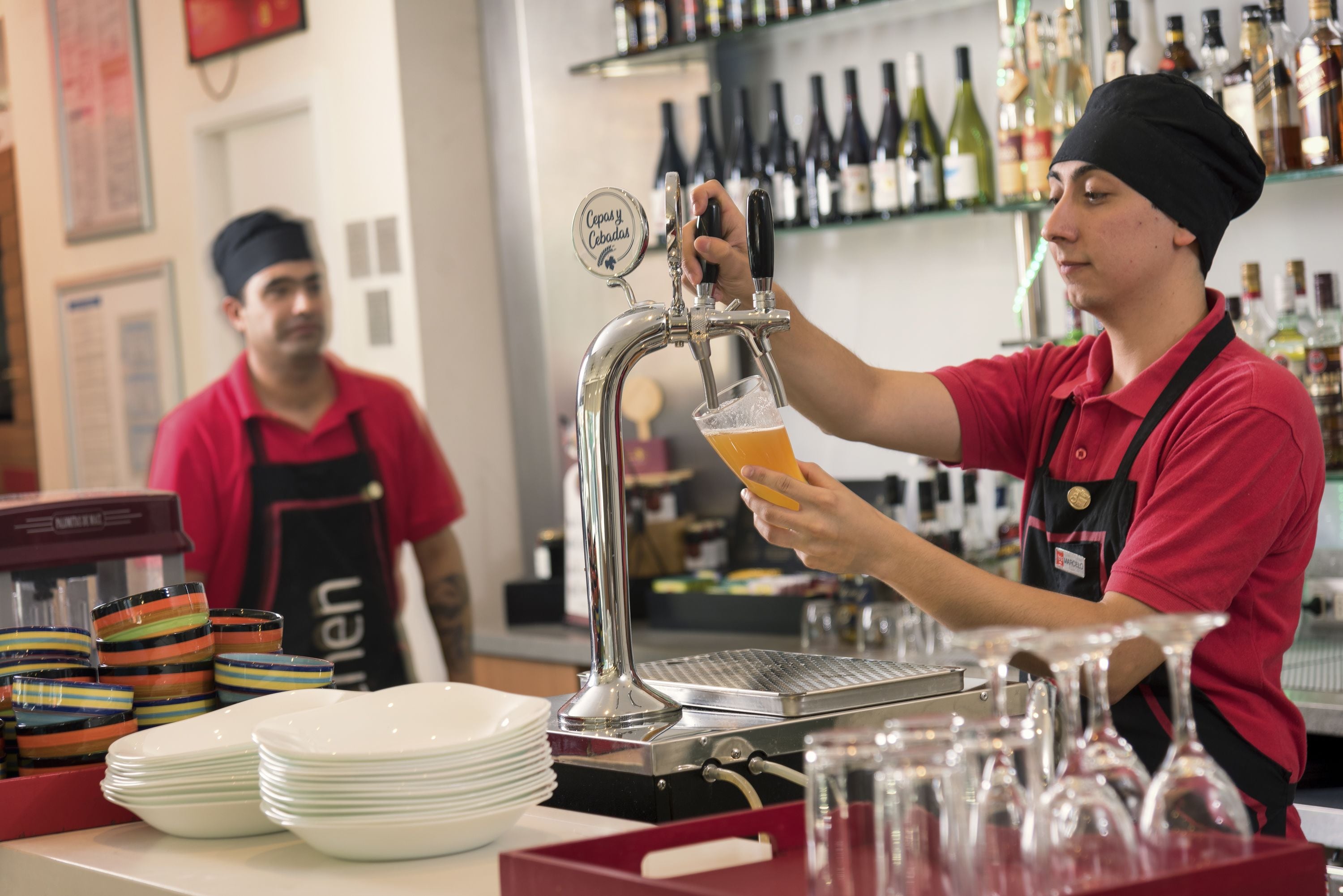 Un barista prepara una bebida en una máquina de café, con varios vasos de café y una estantería para copas de vino al fondo.