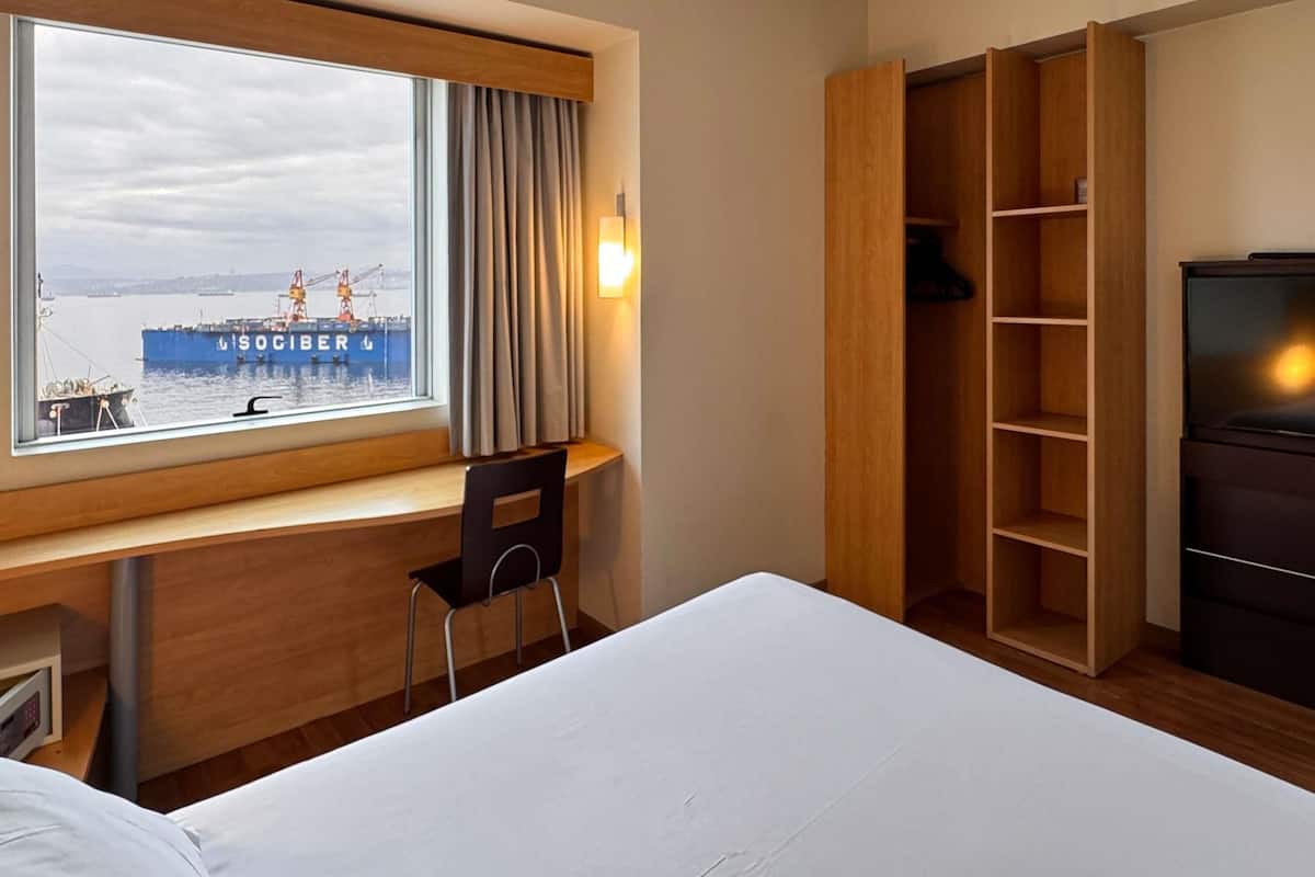 A hotel room with a bed, a desk with a chair, a wardrobe, and a window with a view of a ship and cranes.