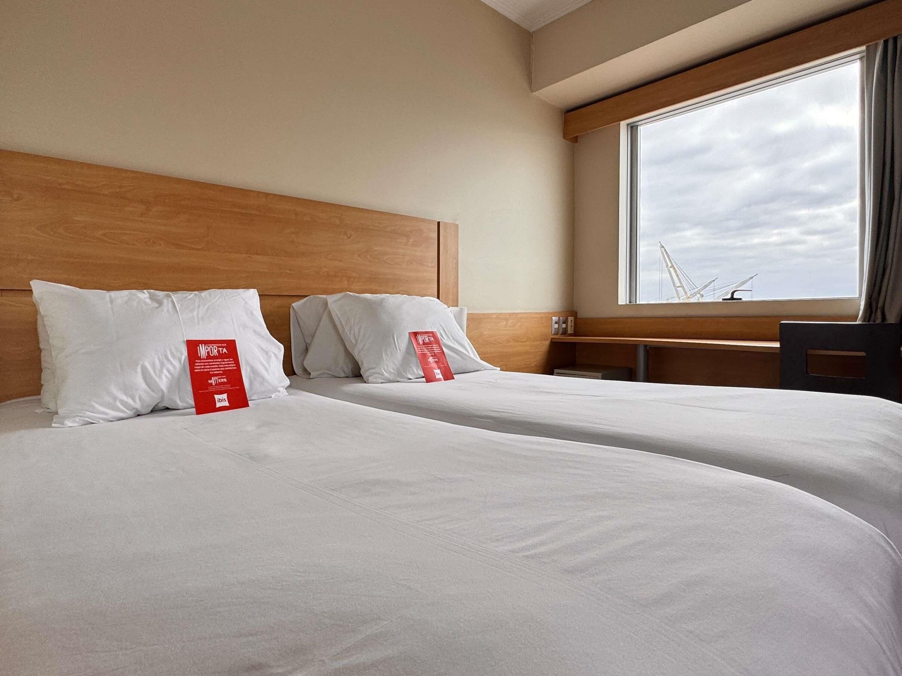 A hotel room with two beds, a wooden headboard, a desk, and a large window with a view of a bridge.
