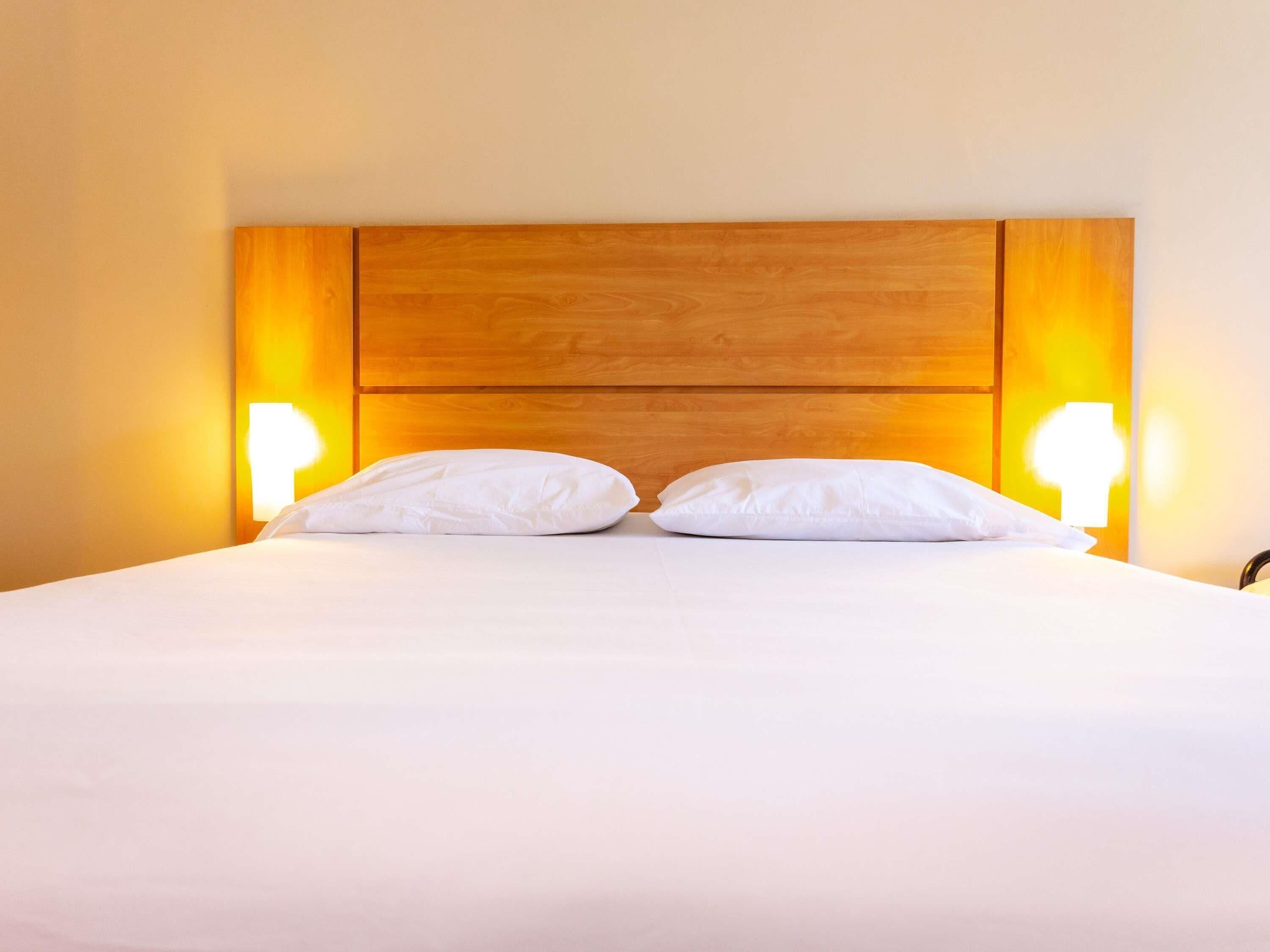 Una cama con cabecera de madera, dos almohadas blancas y dos lámparas fijadas a la pared.