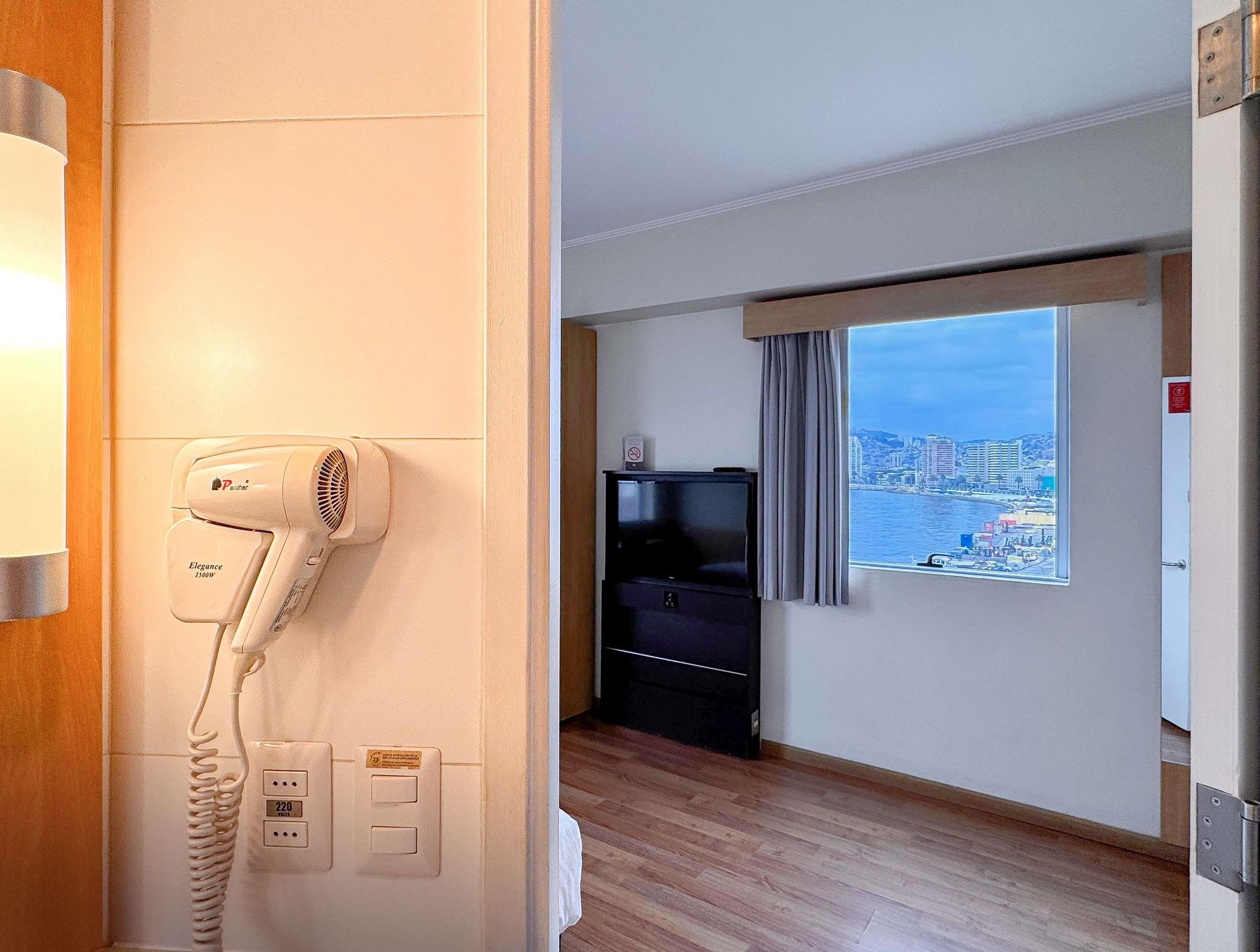Quarto de hotel com secador de cabelo, televisão, janela com vista para a cidade e piso de madeira.