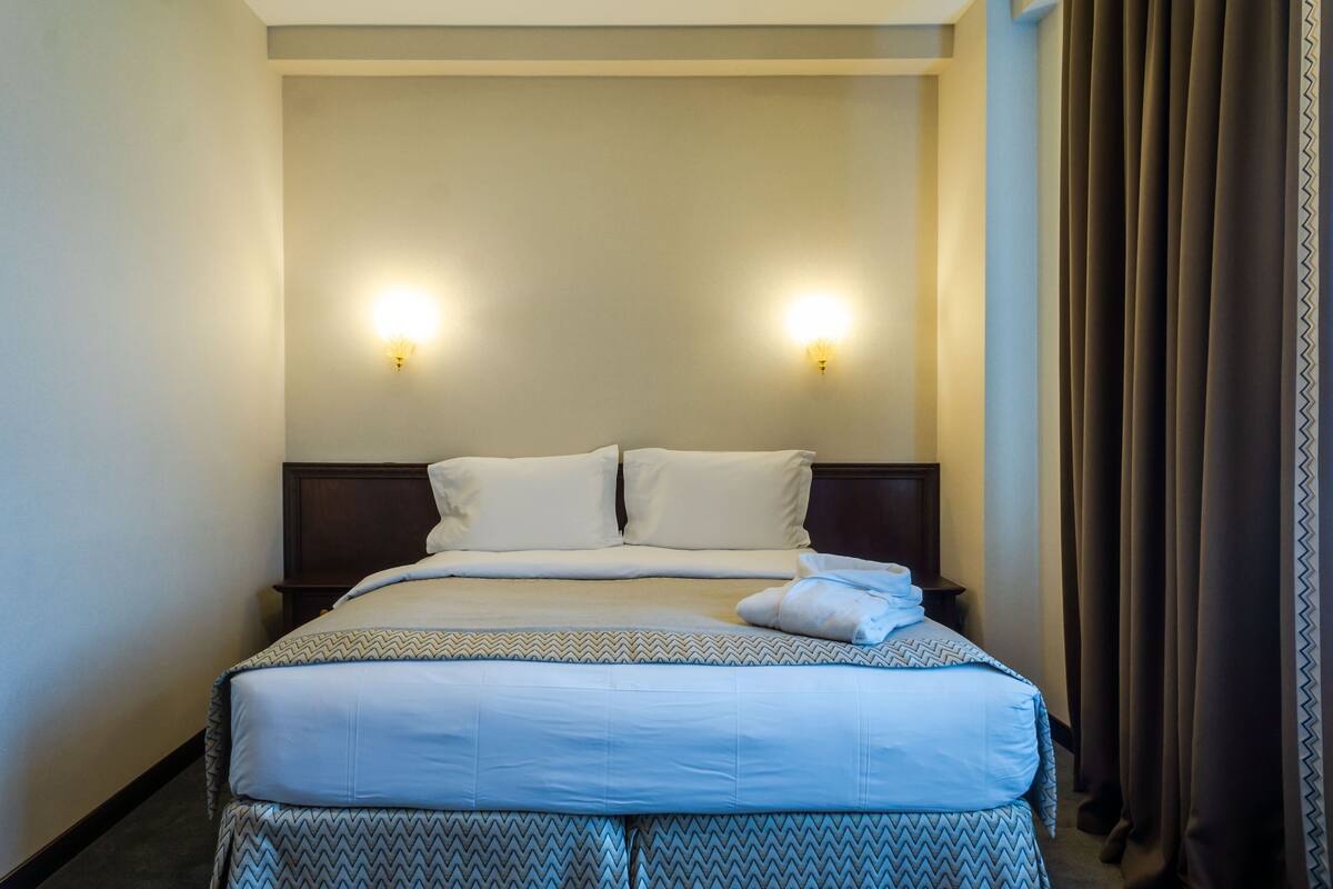 Een hotelkamer met een netjes opgemaakt bed, twee wandlampen en een donker gordijn voor het raam.