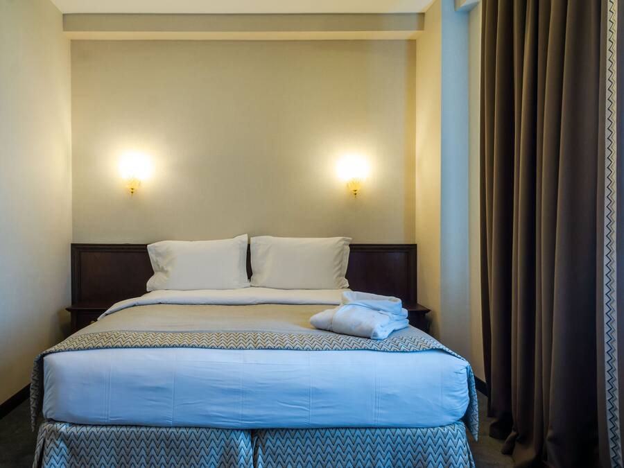A hotel room with a neatly made bed, two wall-mounted lights, and a dark curtain on the window.