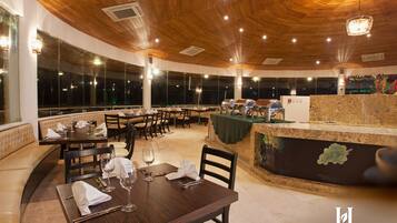 Un restaurante con techo de madera y ventanas amplias, mesas dispuestas con copas de vino y servilletas, y una barra con una exposición de frutas.