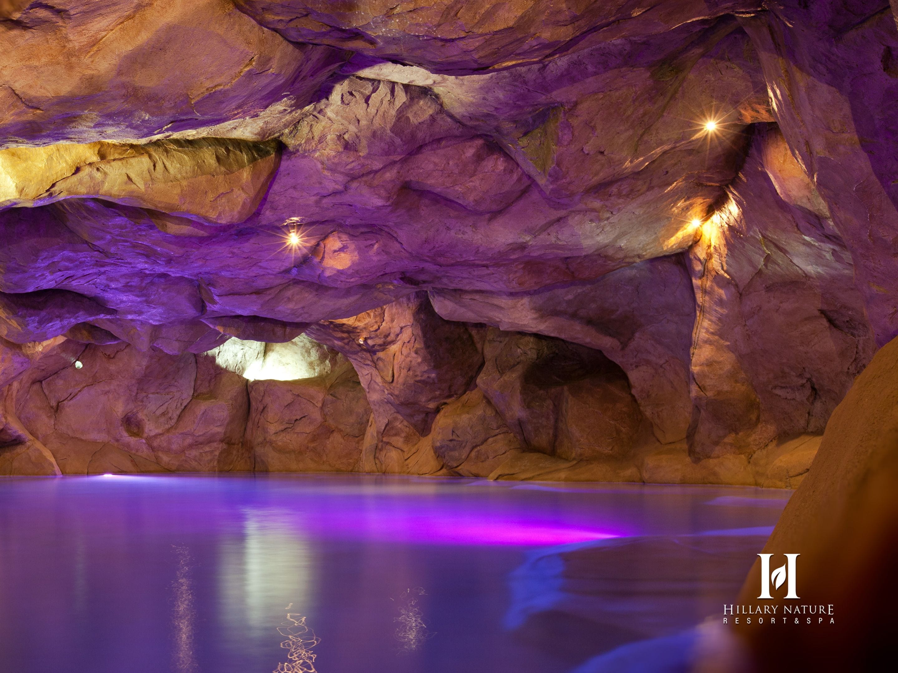 Interior de uma caverna com uma piscina refletindo iluminação roxa, rodeada por formações rochosas.