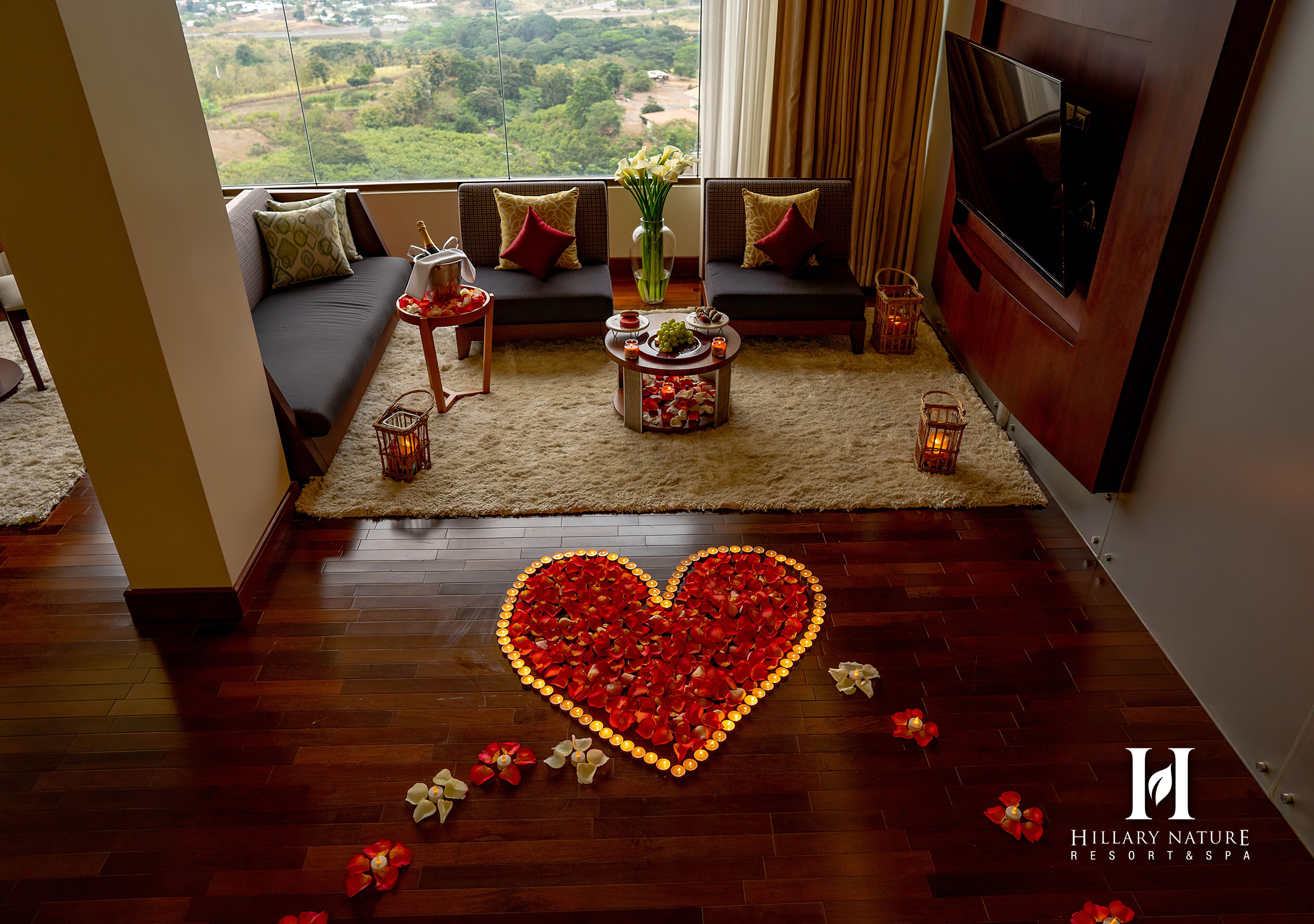 A living room with a heart-shaped arrangement of red flowers on the floor, a sofa with cushions, a small table with a vase and candles, and a TV mounted on the wall.