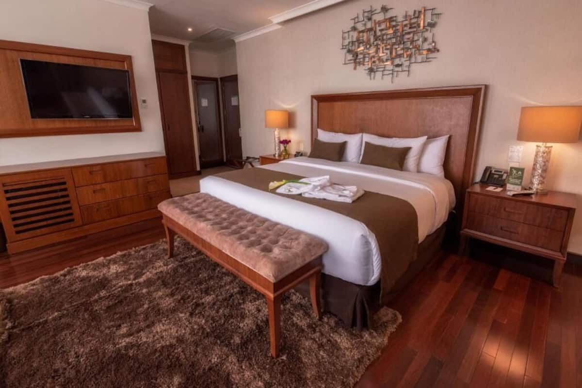 A hotel room with a large bed, a flat-screen TV mounted on the wall, a wooden bench, and a small bedside table with a lamp.