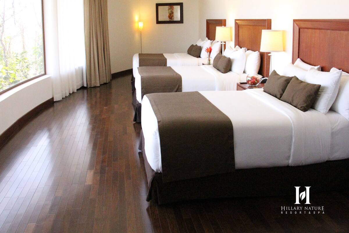 A hotel room with two beds, wooden flooring, a large window with curtains, a wall-mounted lamp, and a framed picture.