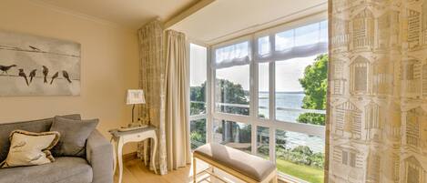 A living room with a grey sofa, a bench, a table with a lamp, and a large window with a view of a body of water and greenery.