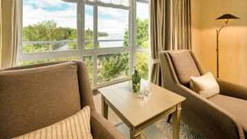 A room with a view of a lake, a sofa, a chair, a table with a glass and a bottle, and a floor lamp.
