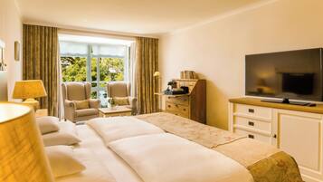 A hotel room with a large bed, a television, a desk with a lamp, a chair, and a view of greenery outside.