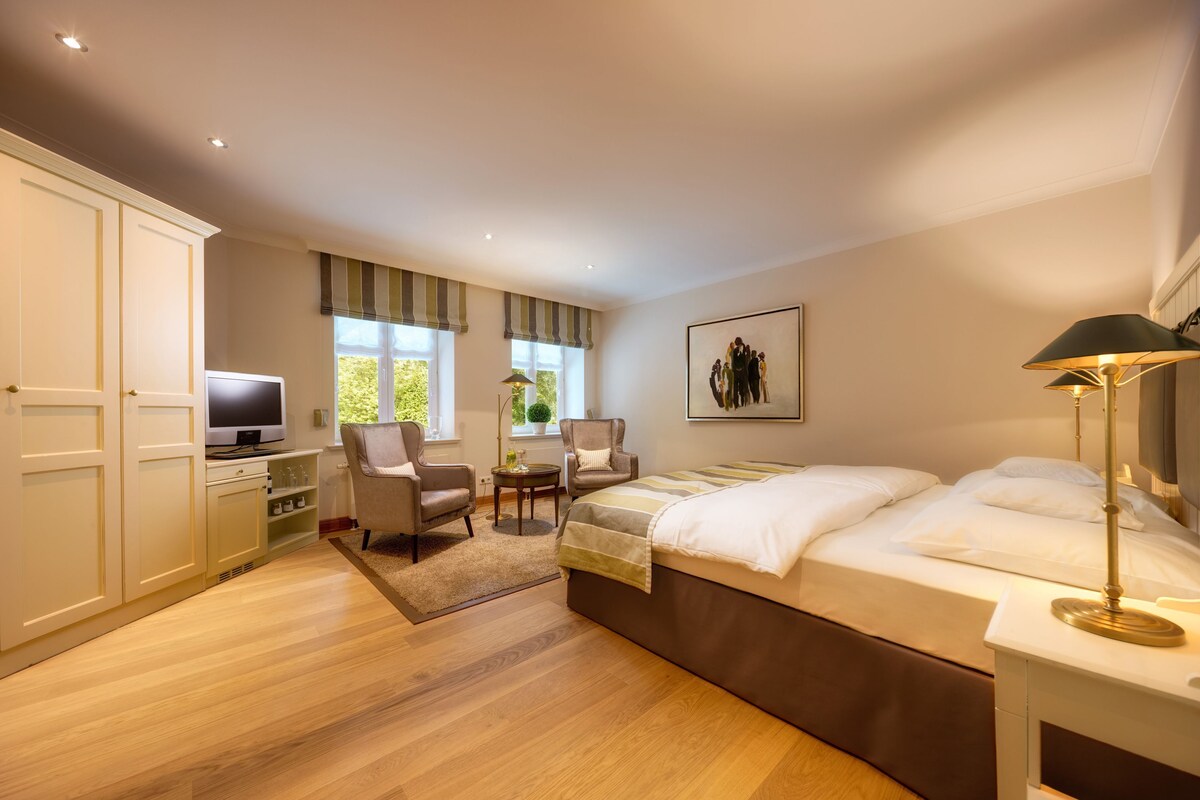 A bedroom with a bed, two chairs, a TV, a desk, and a window with a view.