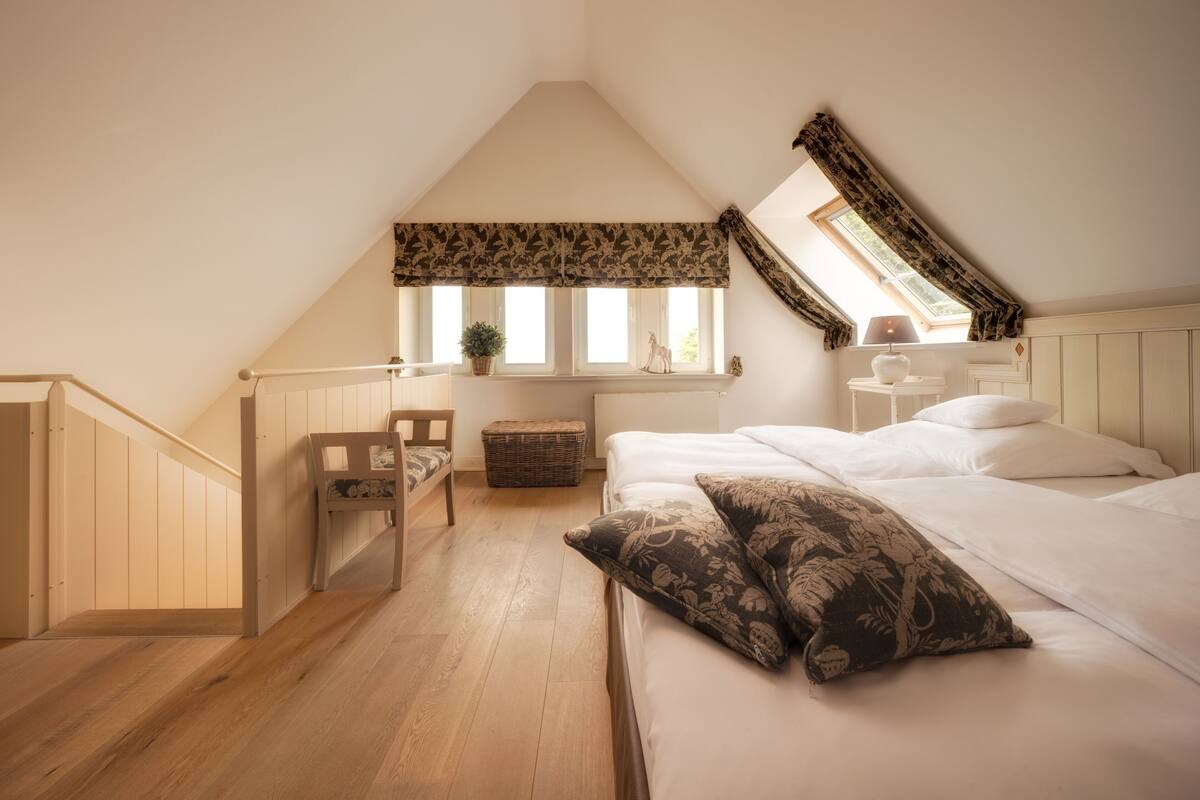 A bedroom with a sloped ceiling, a bed with white bedding, a small table with a lamp, and a window with a view of the outdoors.