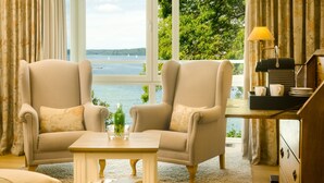 Una habitación con dos sillones mirando hacia una ventana que ofrece vistas a un lago y árboles. Hay una pequeña mesa con libros y una lámpara.
